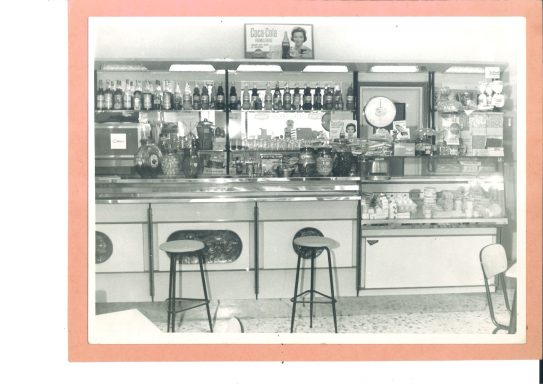 Vintage black-and-white photo of a bar interior with shelves of bottles and stools.